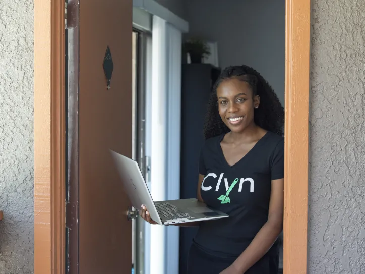 A woman stands by her front door and holds her laptop 