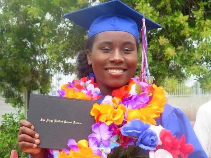 A woman holds her diploma and smiles as she graduates in her graduation gown