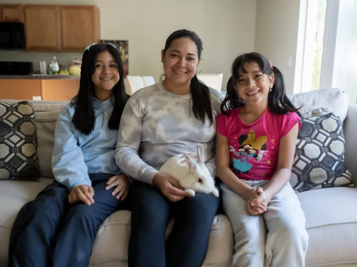 A woman, her two daughters, and their pet rabbit sit on their sofa and smile for a photo 