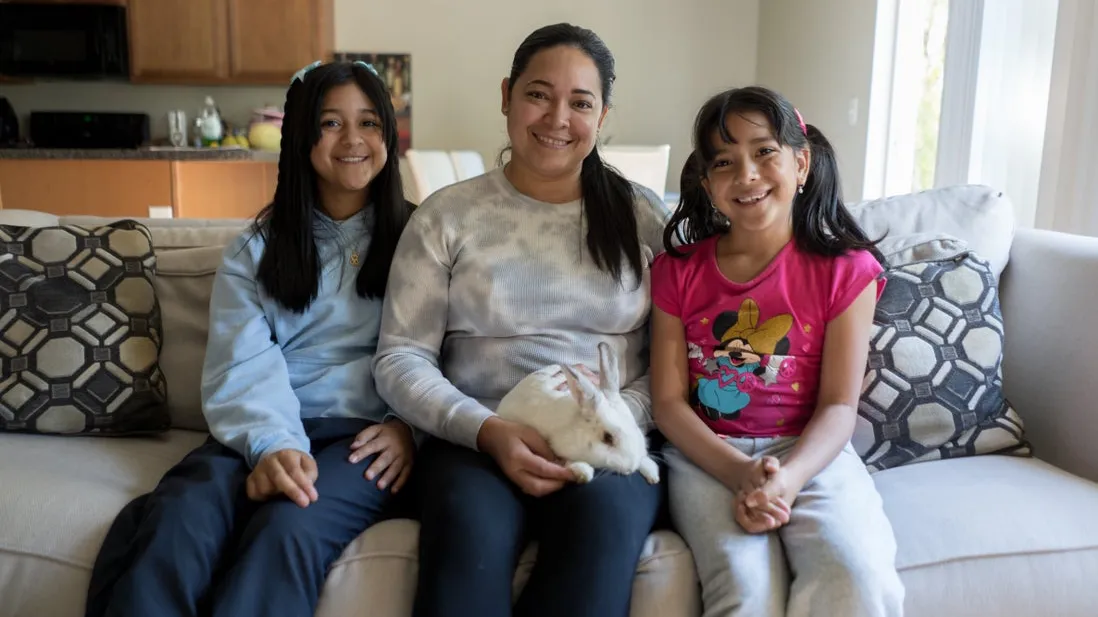 A woman, her two daughters, and their pet rabbit sit on their sofa and smile for a photo 