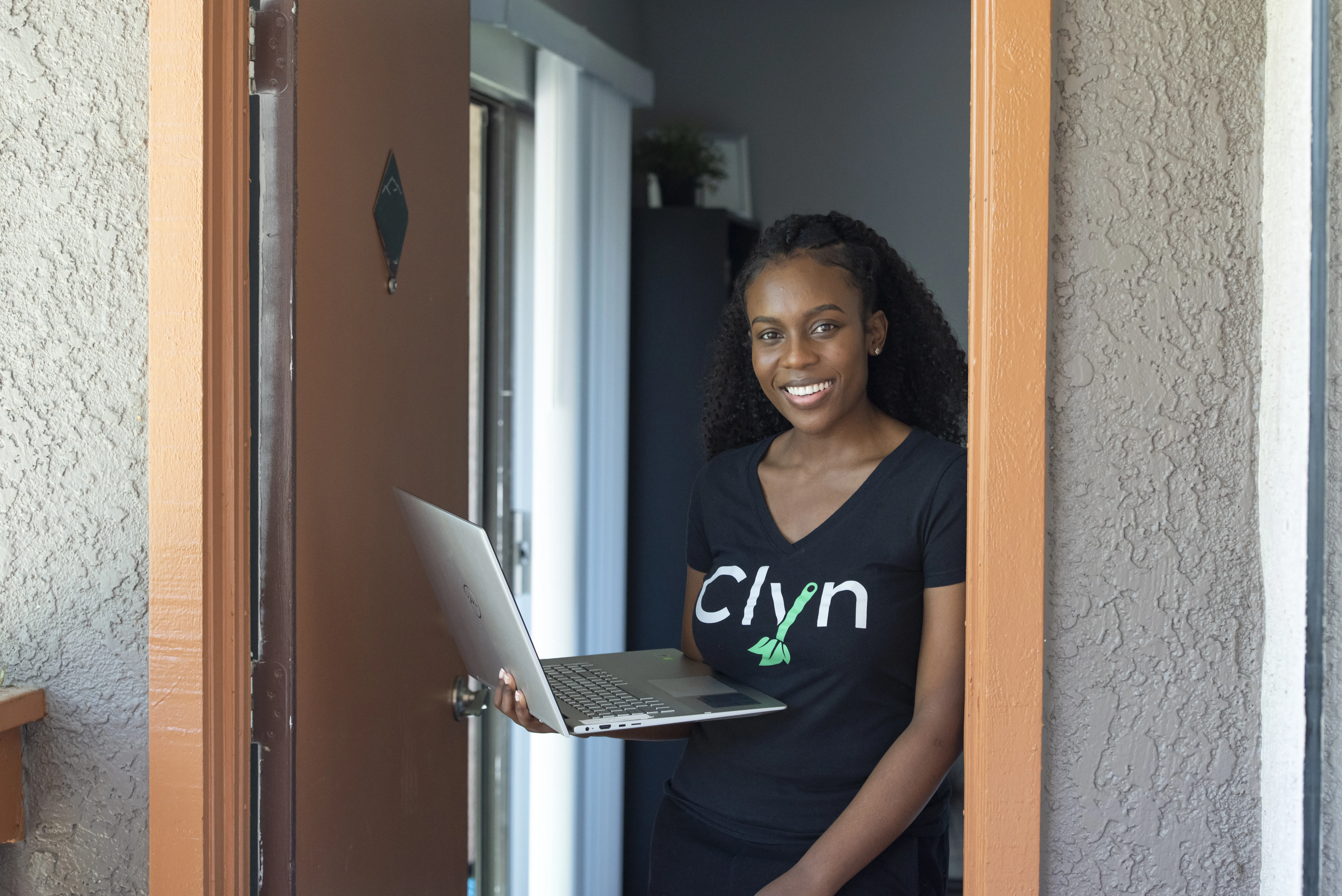 A woman stands by her front door and holds her laptop 