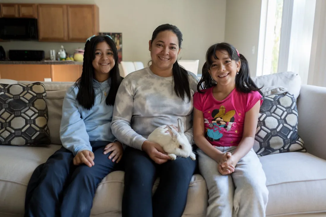 A woman, her two daughters, and their pet rabbit sit on their sofa and smile for a photo 